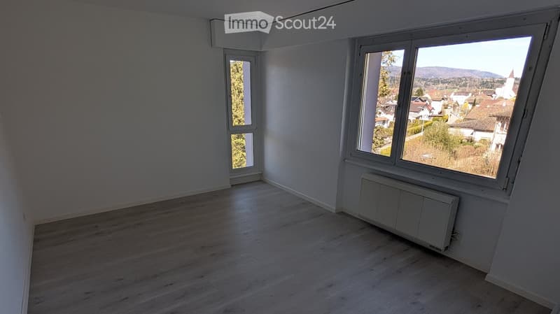 Empty room with light wooden flooring, white walls, and large windows offering a view of the town and surrounding landscape.