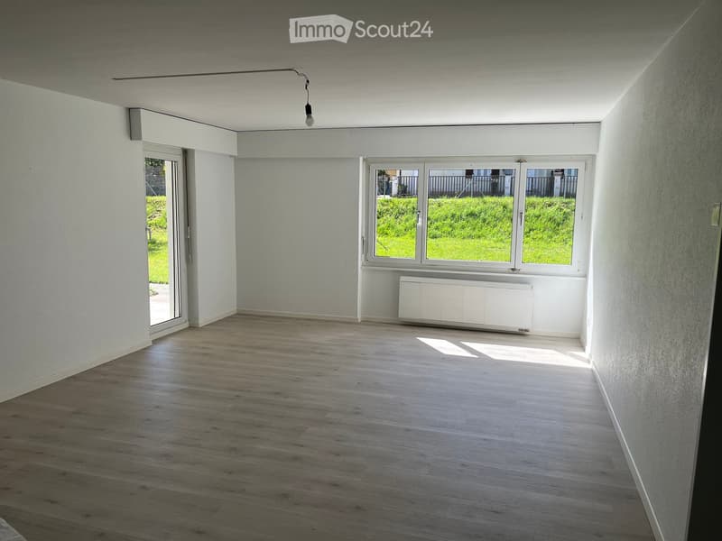 Empty room with light wooden flooring, white walls, large windows, and a radiator. No furniture or equipment present.