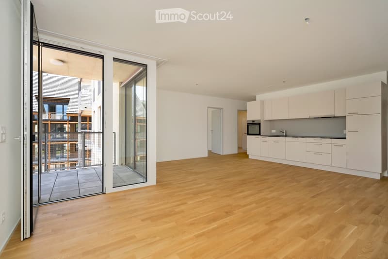 Open-plan living and kitchen area with light wooden flooring, white kitchen cabinets, and a built-in oven. Large sliding glass doors lead to a balcony.
