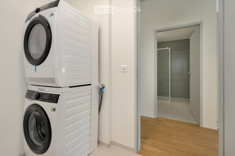 A compact laundry area with a stacked washer and dryer unit against a white wall, adjacent to a wooden-floored hallway leading to a tiled bathroom.