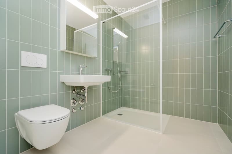 Modern bathroom with green tiled walls, white floor, toilet, sink, mirror, and glass-enclosed shower area.
