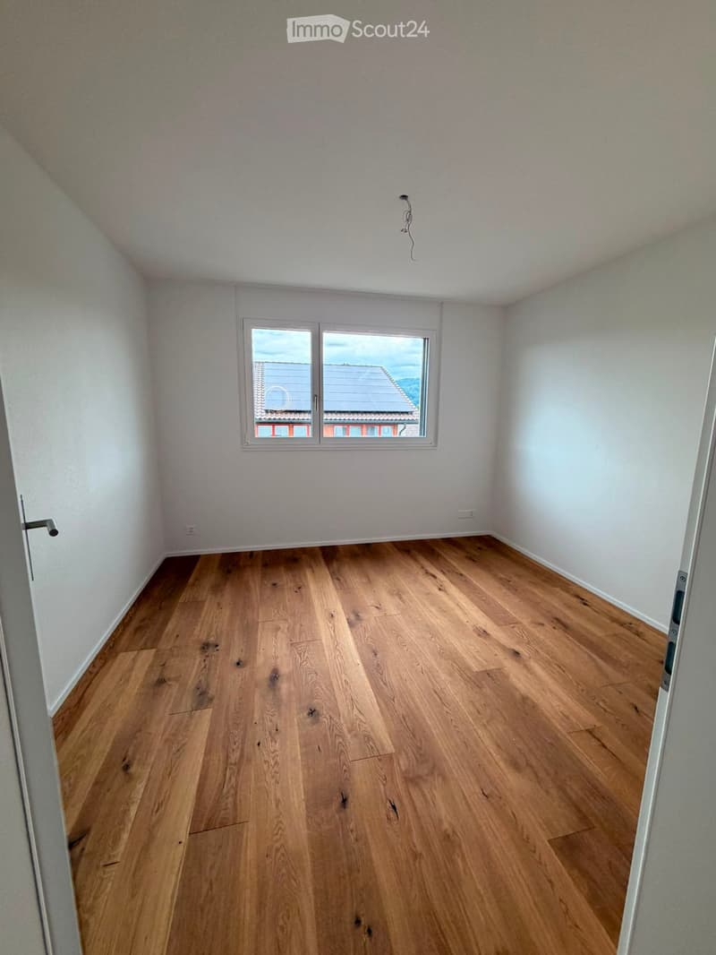 Empty room with wooden flooring, white walls, and a window showing a view of a neighboring building roof.