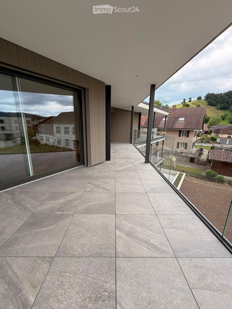 Modern balcony with large glass railings, concrete tiled floor, and expansive view of surrounding houses and landscape.