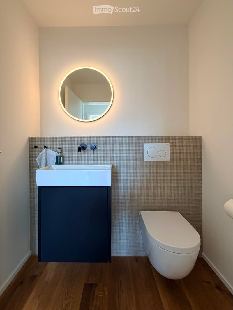 Modern bathroom with a white sink, dark cabinet, round illuminated mirror, and a toilet. The walls are tiled with a textured pattern, and the floor is wooden.