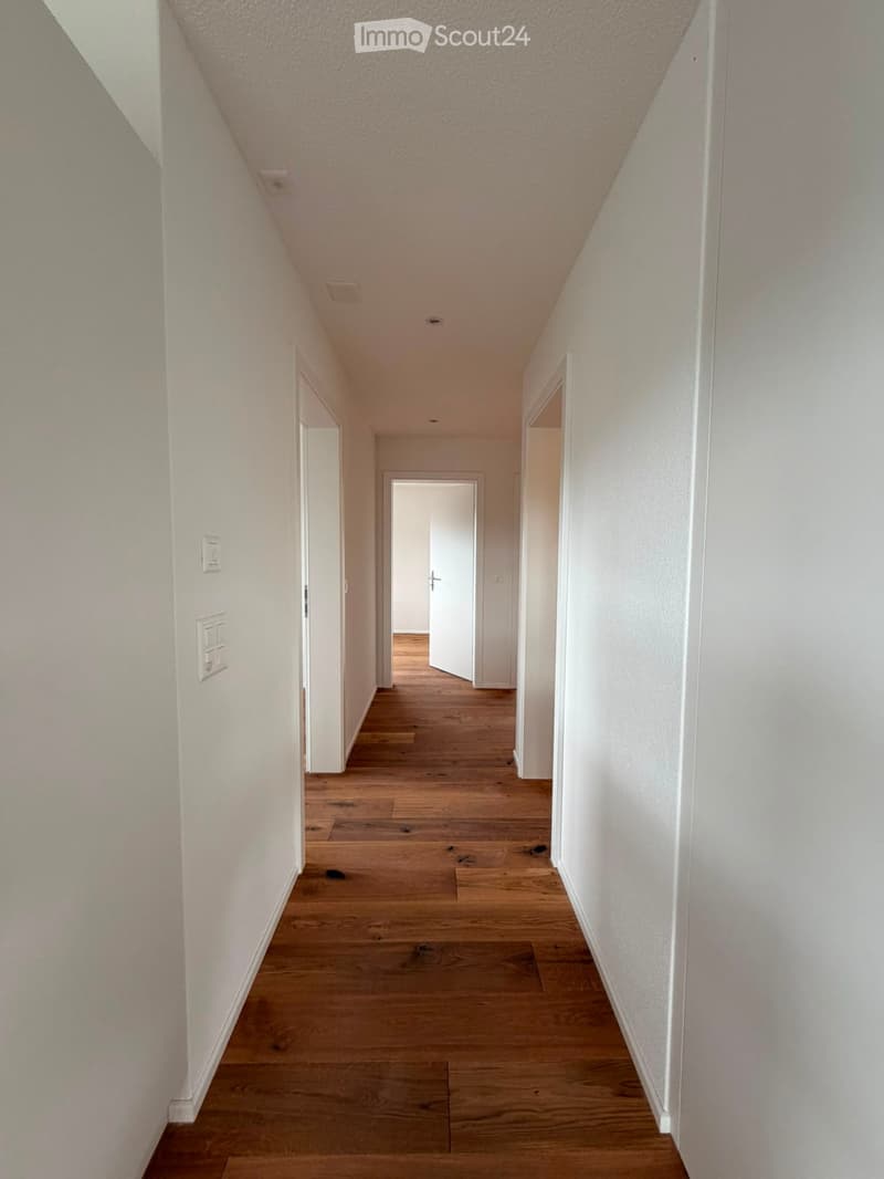 A narrow hallway with wooden flooring and white walls, featuring several doors and light switches on the walls.