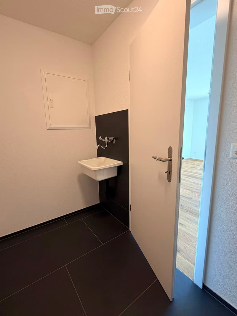 Modern bathroom with dark tiled floor, white sink, and black accent wall. Open door leads to another room with wooden flooring.