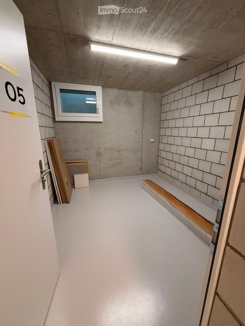 Empty room with white tiled walls, concrete ceiling, and a small window. Wooden plank and small wooden stool are placed against the wall.