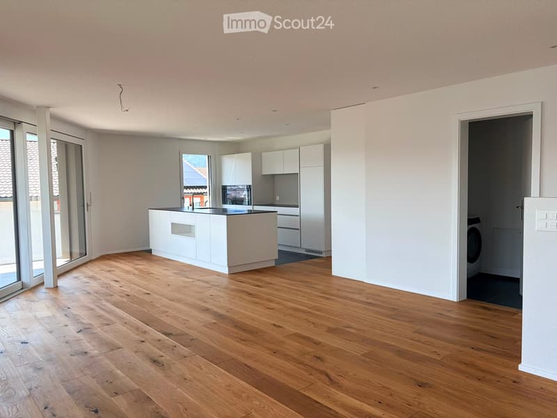 Open-plan living and kitchen area with light wooden flooring, white kitchen cabinets, a central island, and a washing machine in the corner.