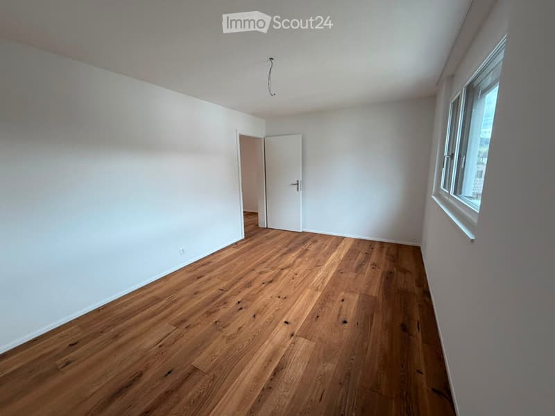 Empty room with wooden flooring, white walls, and a window on one side. A door is visible leading to another space.