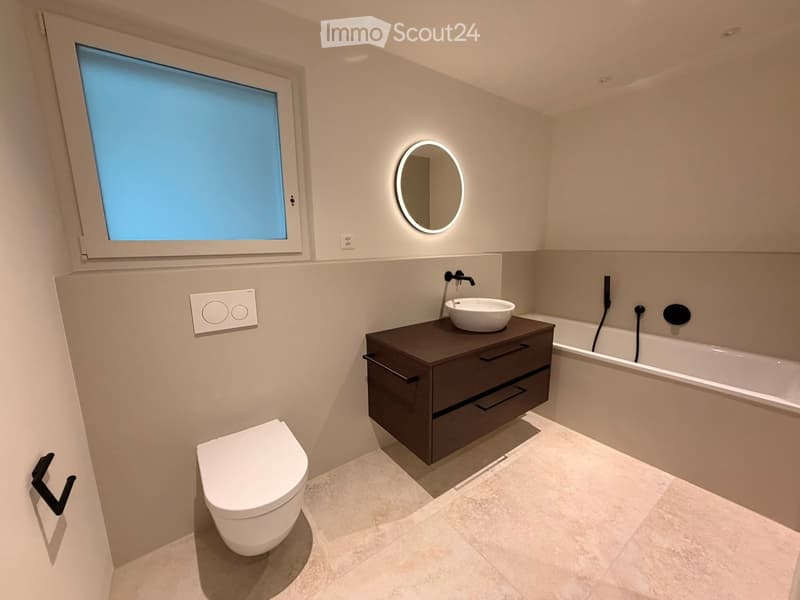 Modern bathroom with a white toilet, dark vanity cabinet with a sink, a bathtub, and a round illuminated mirror above the sink. The room features light-colored tiles and a large window.