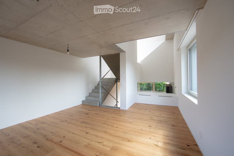 Empty room with wooden flooring, white walls, and a staircase leading upwards. Natural light streams in from large windows, and the ceiling features exposed wooden beams.