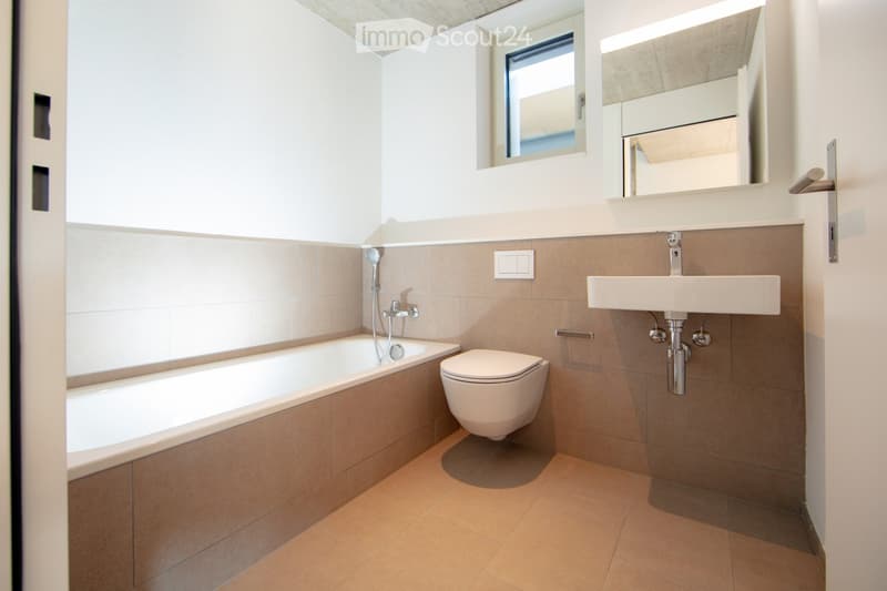 Modern bathroom with a bathtub, toilet, sink, and mirror. Features tiled walls and floor, a window, and a sleek design.
