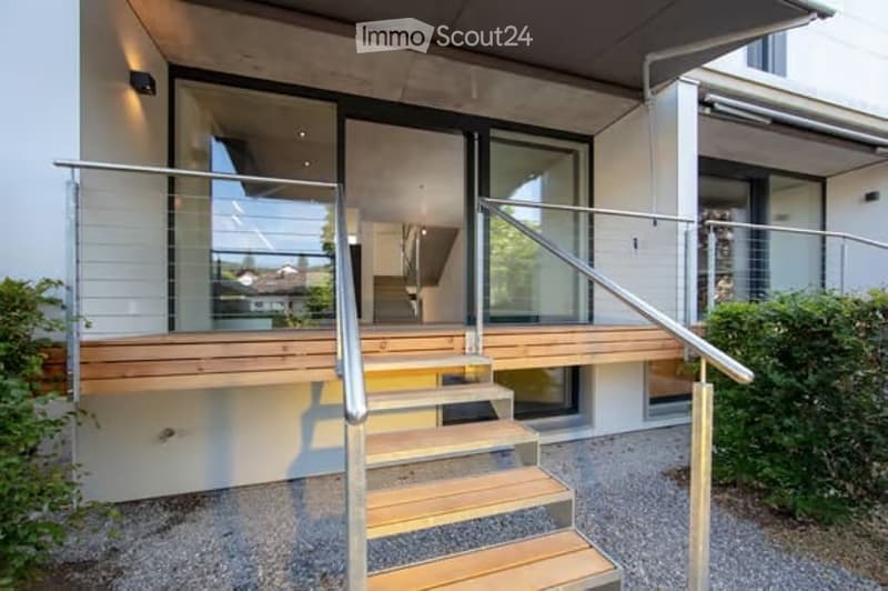 Modern house entrance with wooden steps, metal railing, and large glass doors leading to the interior.