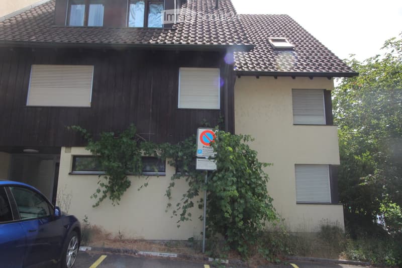 Two-story house with a tiled roof, beige and dark brown exterior, multiple windows with shutters, and a small garden area with climbing plants.