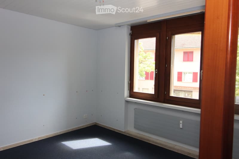 Empty room with white walls, wooden window frame, radiator, and grey carpeted floor.