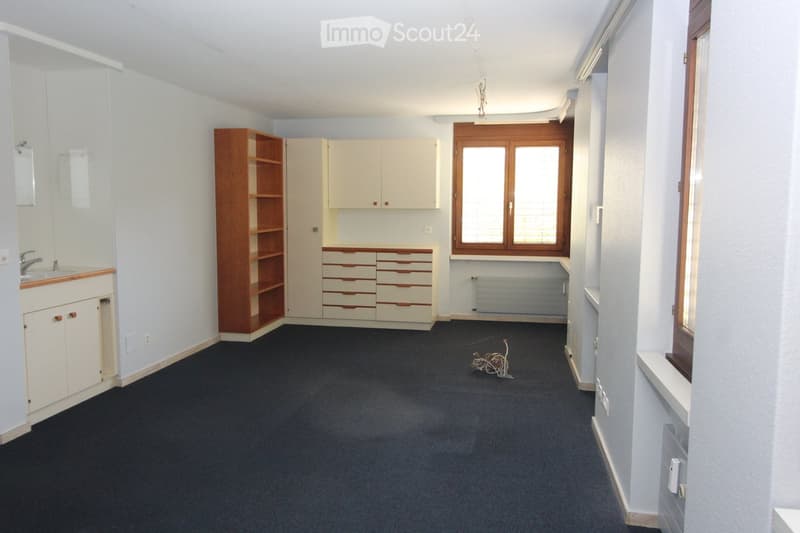 A small, furnished room with a sink, cabinets, a wooden shelf, and a radiator. The room has light-colored walls, a dark carpeted floor, and a window with wooden frames.