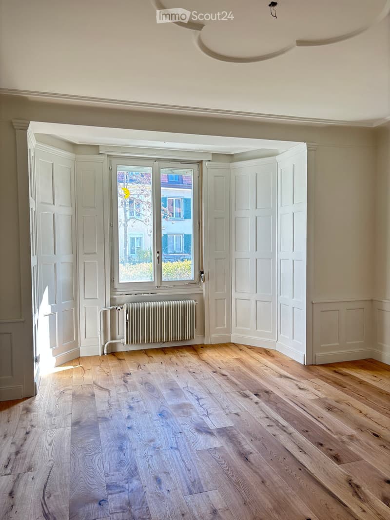 Empty room with light wooden flooring, white walls, large window with view of the street, radiator, and white panel doors.