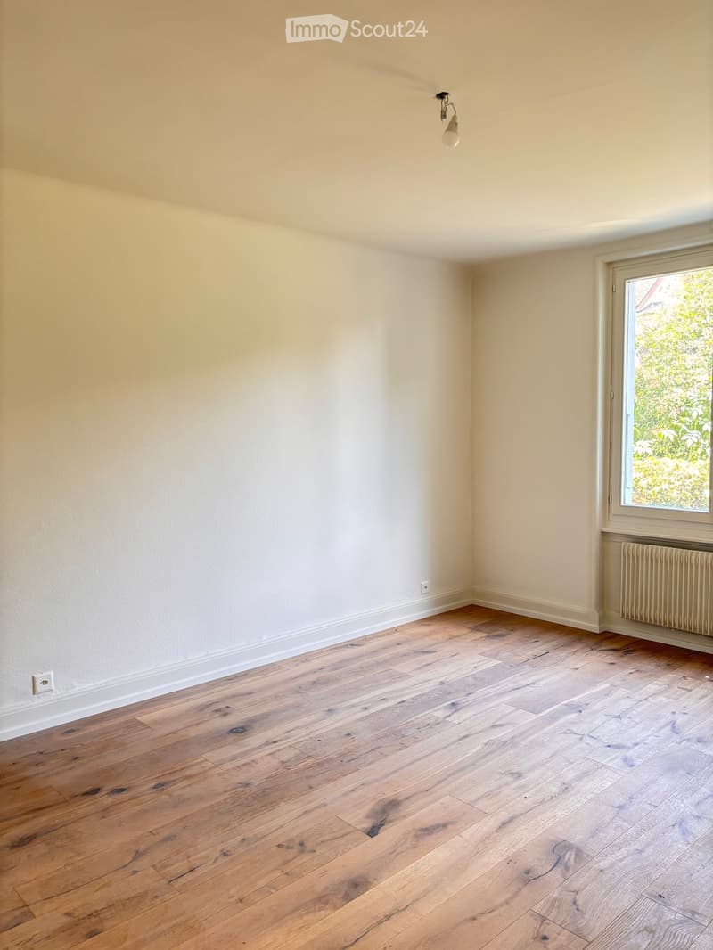 Empty room with light wooden flooring, white walls, and a window with a view of greenery. A radiator is visible under the window, and a single light fixture hangs from the ceiling.