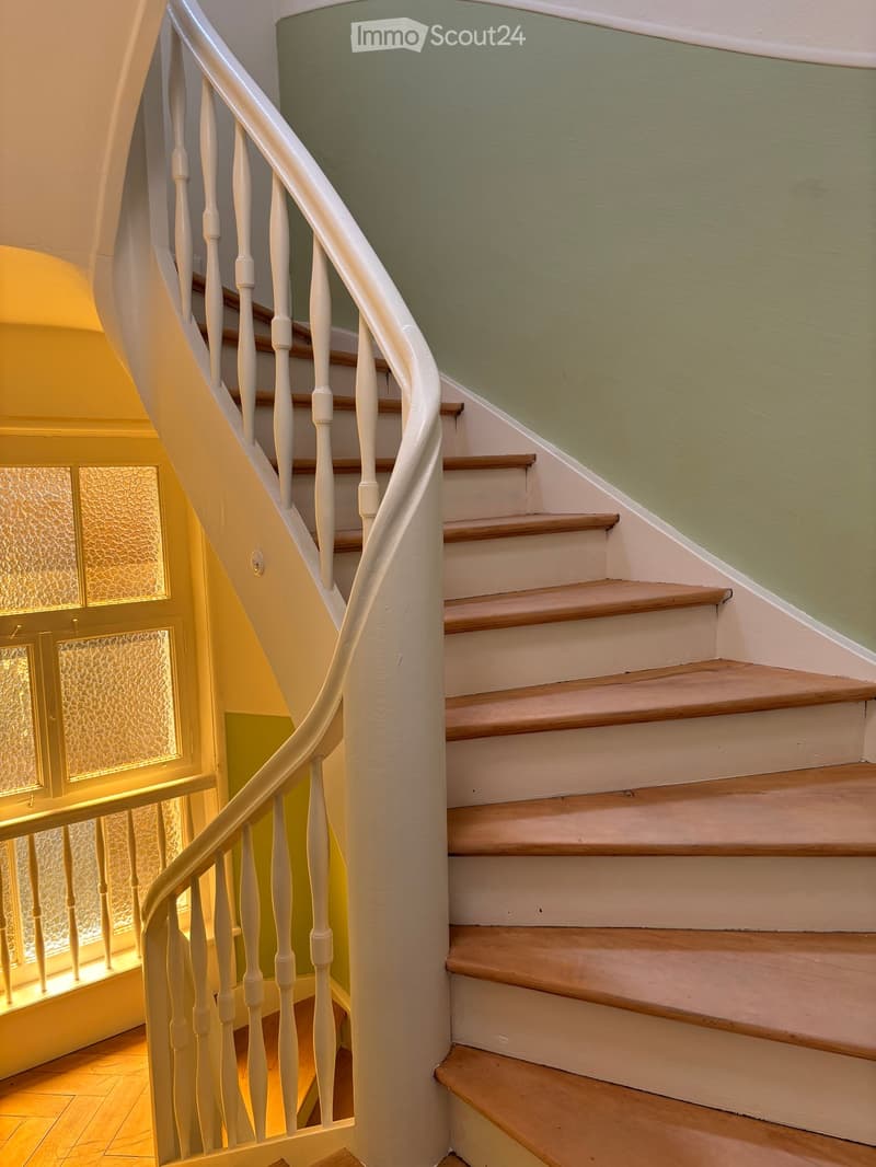 Spiral staircase with wooden steps and white balustrade, leading to an upper level. The walls are painted in a light green color, and the floor is tiled.