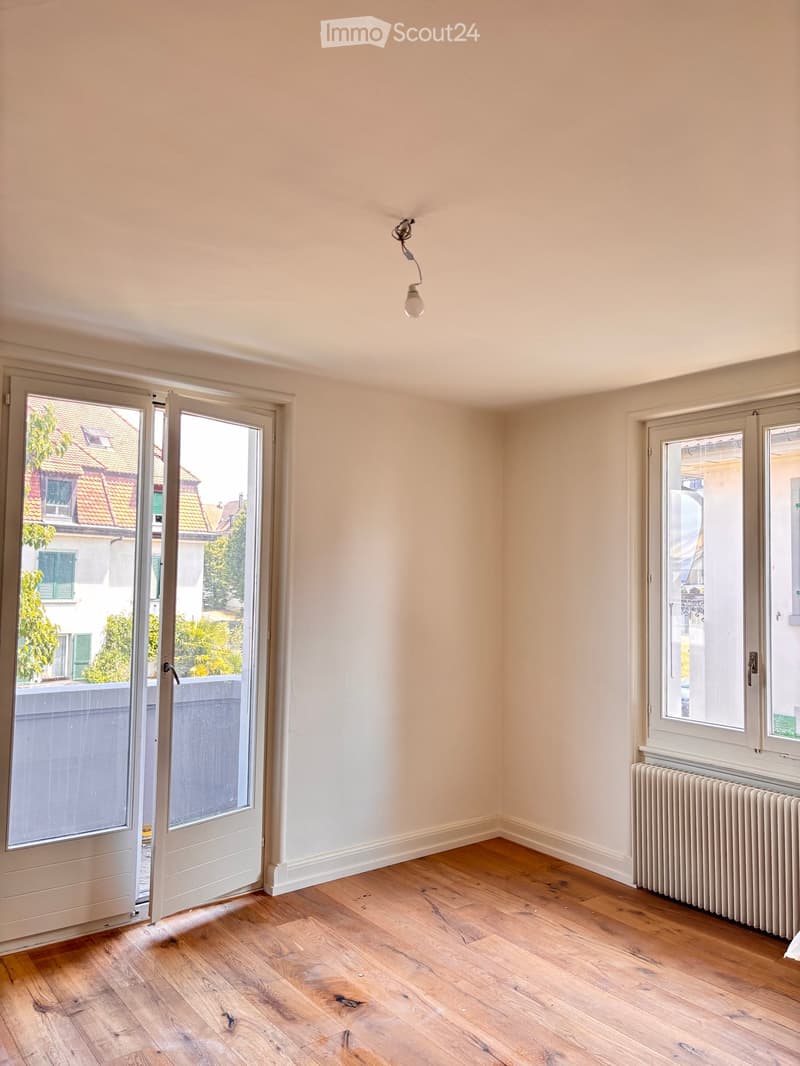Empty room with wooden flooring, white walls, and large windows. A radiator is visible under one window, and a ceiling light fixture is present.