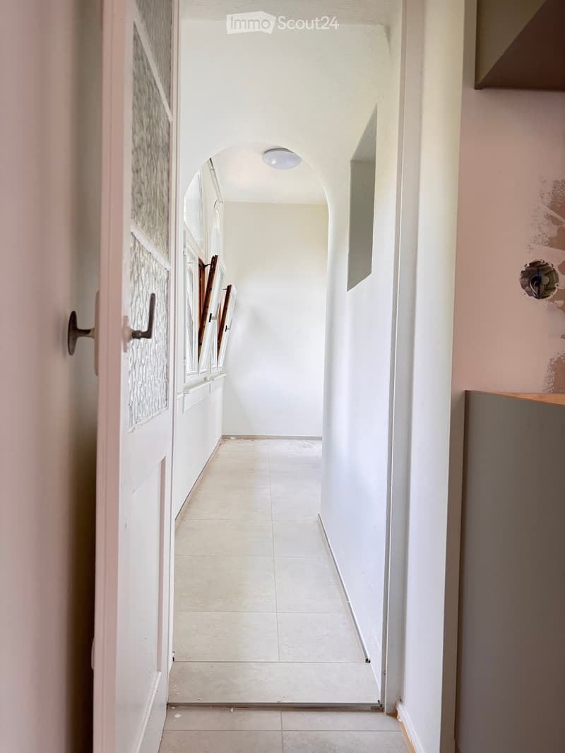 A narrow hallway with white tiled flooring, white walls, and a ceiling light. The hallway features an arched doorway at the end and a partially open door on the left side.