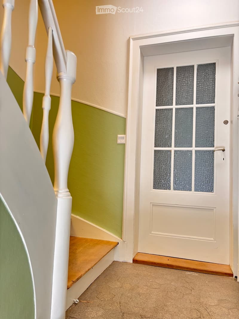 Hallway with a staircase leading upwards, green and beige walls, wooden steps, white balusters, and a frosted glass door at the end.