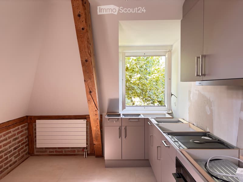 Modern kitchen with light pink cabinets, white countertop, and stainless steel appliances, featuring a large window with a view of greenery.