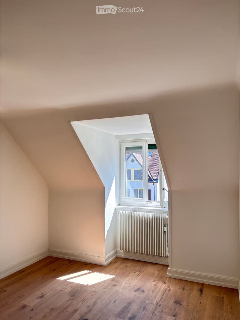 Empty attic room with wooden flooring, slanted ceilings, and a window showing a view of neighboring houses.