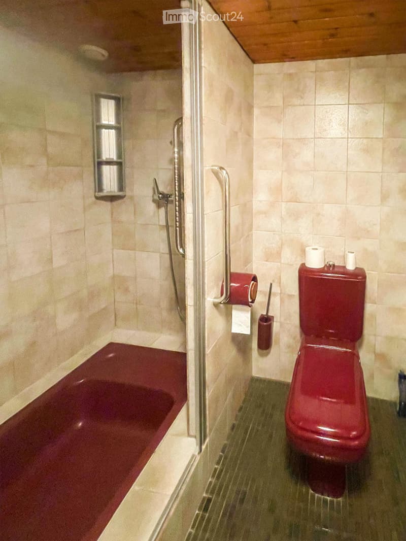 Bathroom with a red bathtub, red toilet, shower area with a glass enclosure, and a toilet paper holder. The walls are tiled in light beige, and the floor is green tiled.
