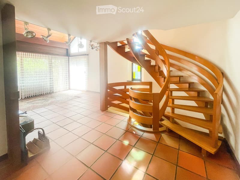 Spacious hallway with a modern wooden spiral staircase, tiled floor, large windows with blinds, and recessed ceiling lights.