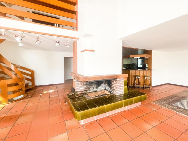 Open-plan living and dining area with a central brick fireplace, wooden staircase, and a small kitchen area with cabinets and stools.