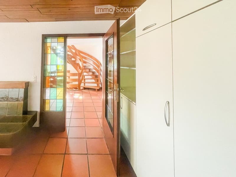 A hallway with a wooden ceiling, tiled floor, and a spiral staircase leading to another room. The hallway features white cabinets and a glass door with colorful stained glass panels.
