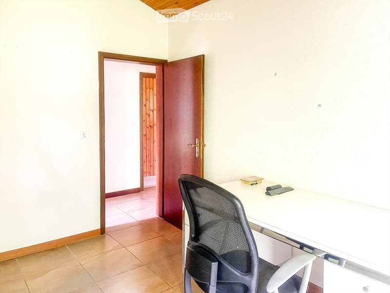 A small, unfurnished room with a tiled floor, white walls, and a wooden door leading to another room. A single chair and a small table are present.