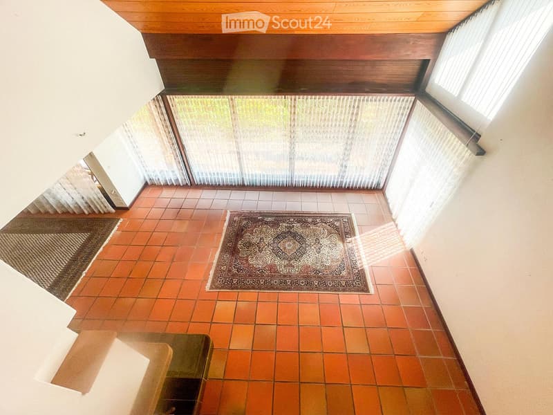 A modern, open-concept living space with terracotta tile flooring, large windows with blinds, and a central patterned rug.