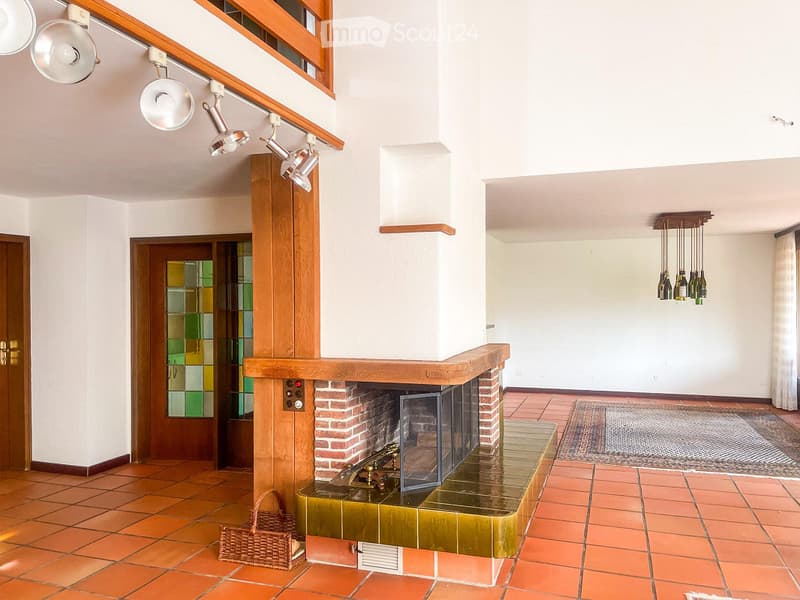 Spacious room with terracotta tiled floor, central brick fireplace with green tiled surround, hanging pendant lights, and a wicker basket on the floor.