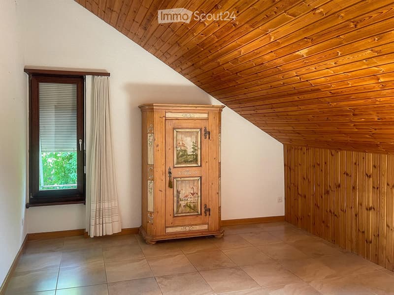 An unfurnished room with wooden ceiling, tiled floor, and a large window with curtains. A wooden cabinet with decorative panels is placed against the wall.