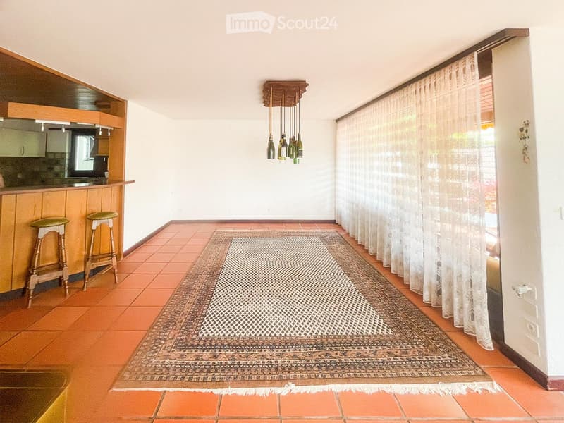 A well-lit room with terracotta tiled flooring, a large patterned rug, two wooden stools, a kitchen counter with a screen, and a wine rack with bottles hanging from the ceiling.