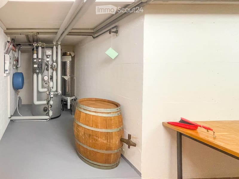 Utility room with a wooden table, red tools, a large wooden wine barrel, and a heating unit with pipes and valves.