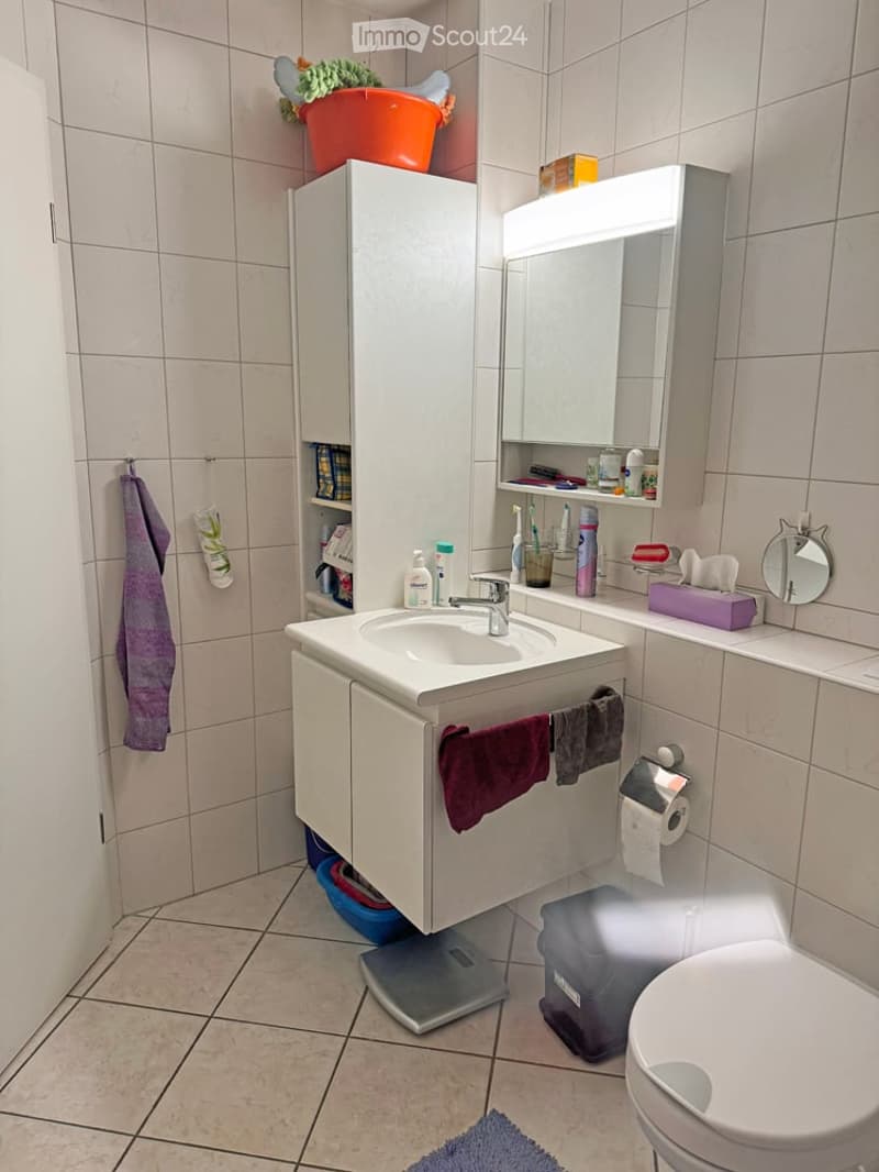 A modern bathroom with a white sink, mirror, toilet, and cabinet. The sink has a chrome faucet and is surrounded by various toiletries. A purple towel hangs on the side of the cabinet, and a small round mirror is placed on the counter. A scale is position