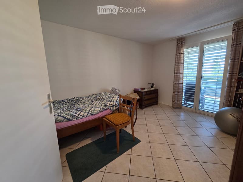 Bedroom with tiled floor, bed with plaid blanket, wooden chair, nightstand, and exercise ball near the window.