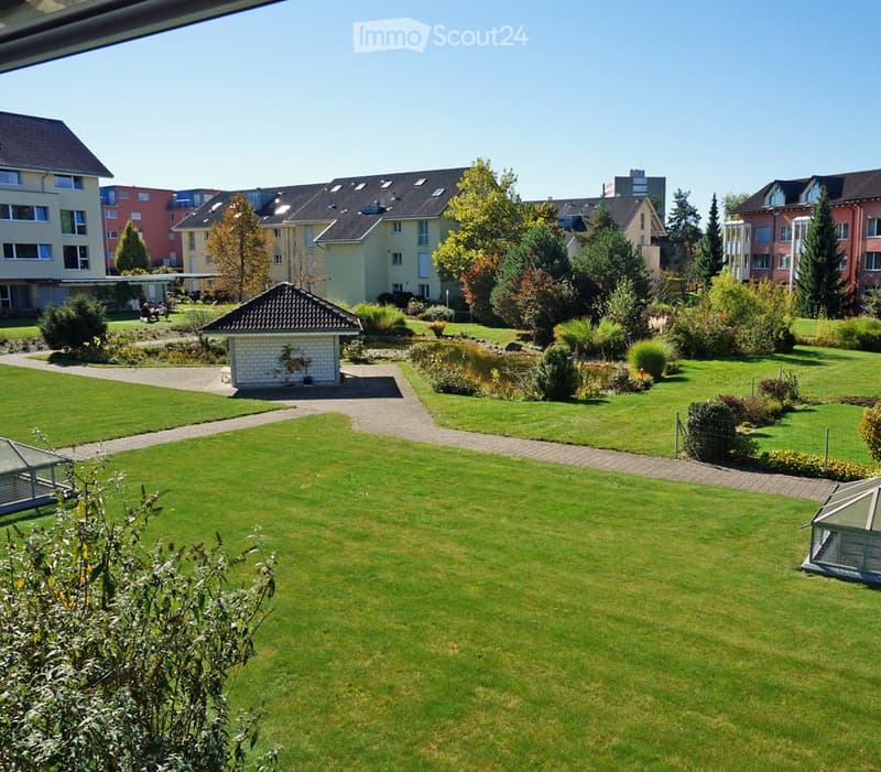 Well-maintained garden area with green lawns, pathways, small structures, and surrounding residential buildings.