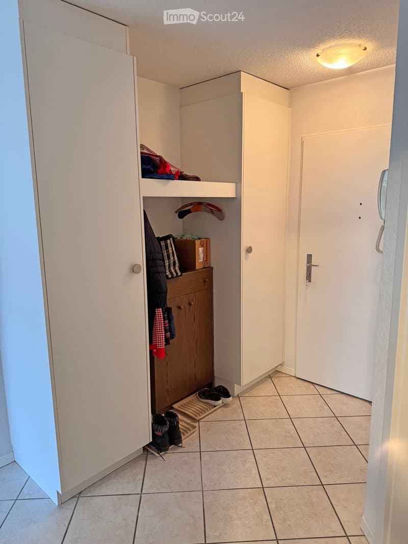 A narrow hallway with tiled flooring, white walls, and built-in storage units. Shoes and clothing are placed near the entrance.