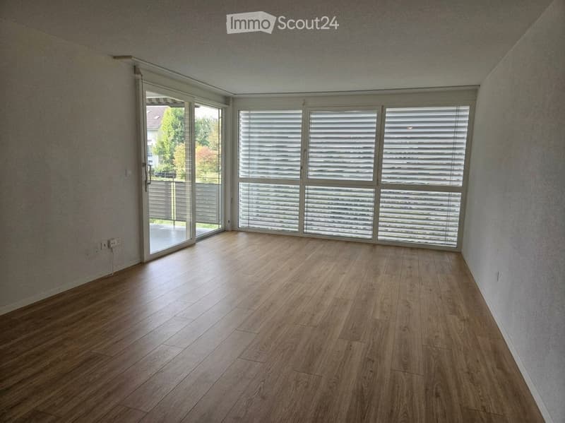Empty room with wooden flooring, large windows with blinds, and a sliding glass door leading to an outdoor area.