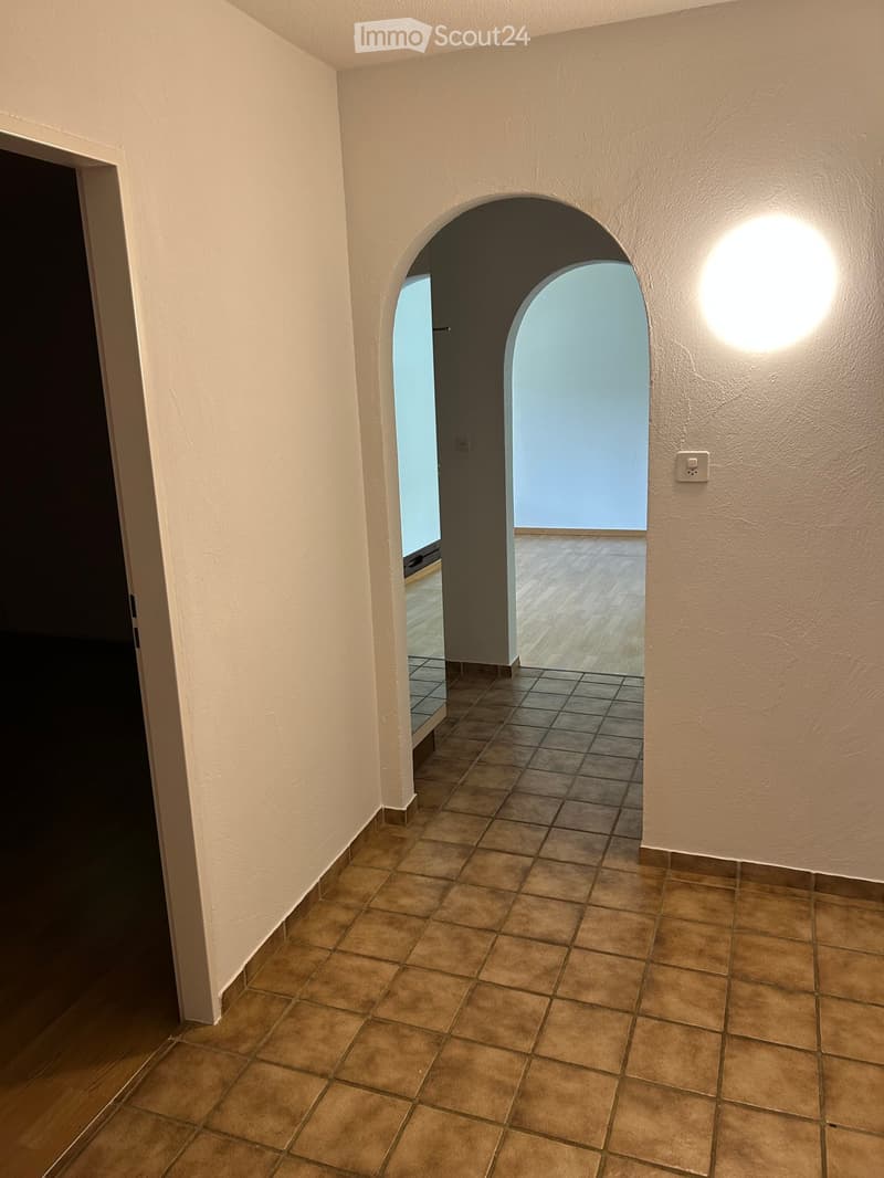Empty hallway with beige tiled floor, white walls, and an arched opening leading to another room with wooden flooring and large windows.