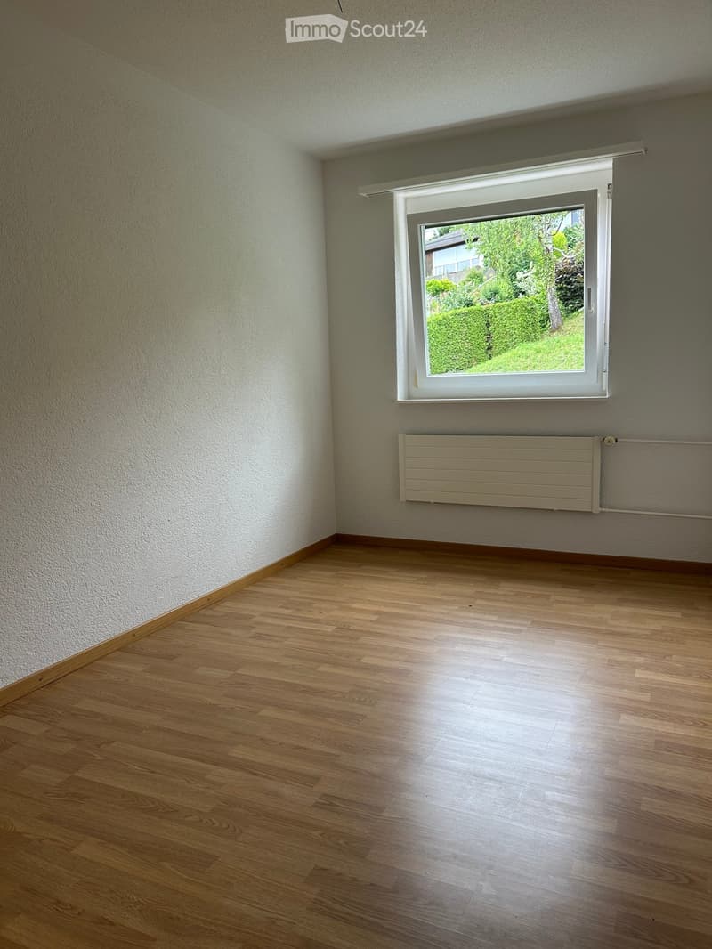 Empty room with wooden flooring, white walls, a large window with white frame, and a radiator below the window.