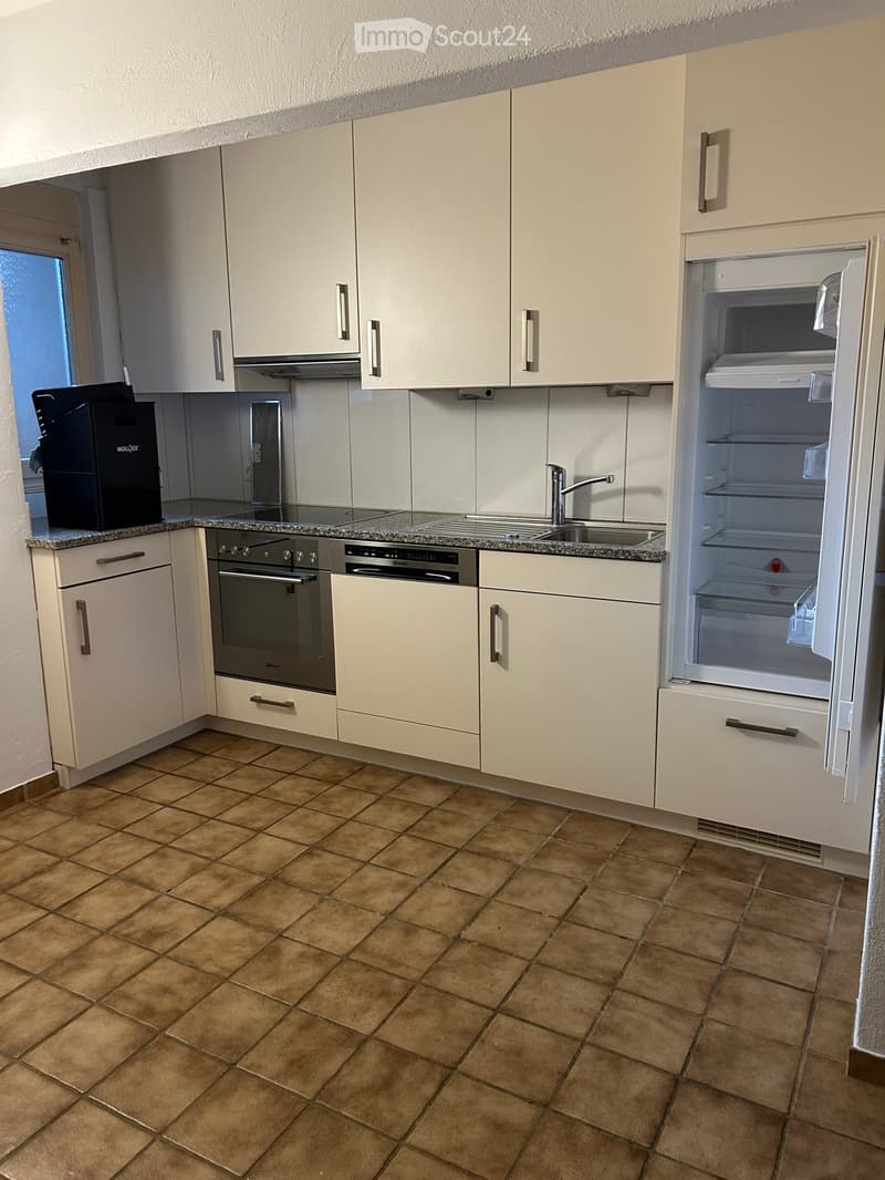 Modern kitchen with light-colored cabinets, granite countertop, stainless steel oven, and open refrigerator. Brown tiled floor.
