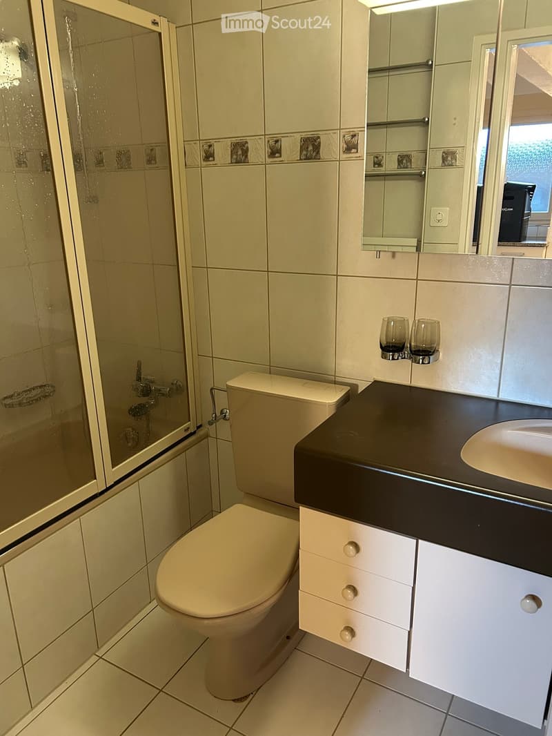 Modern bathroom with white tiles, a toilet, a sink with a black countertop, a mirrored cabinet, and two cups on the countertop.