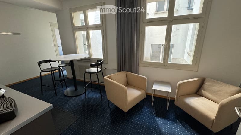 A modern, well-lit room with large windows, a sofa, a chair, a small table, and a round dining table with stools.