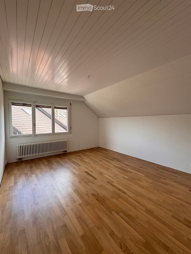 Empty attic room with wooden flooring, white walls, and a slanted wooden ceiling. Three windows offer a view of the roof.