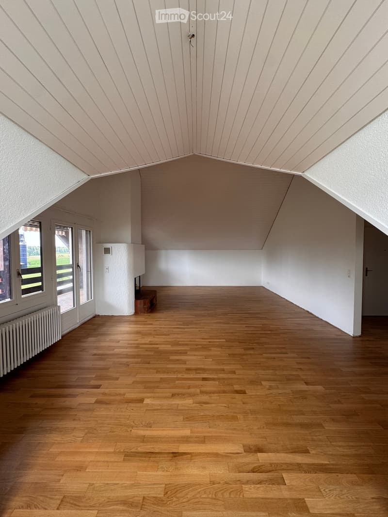 Empty room with wooden flooring, white walls, and a slanted ceiling. Features include a radiator, a small bench, and a window with a view of the outdoors.